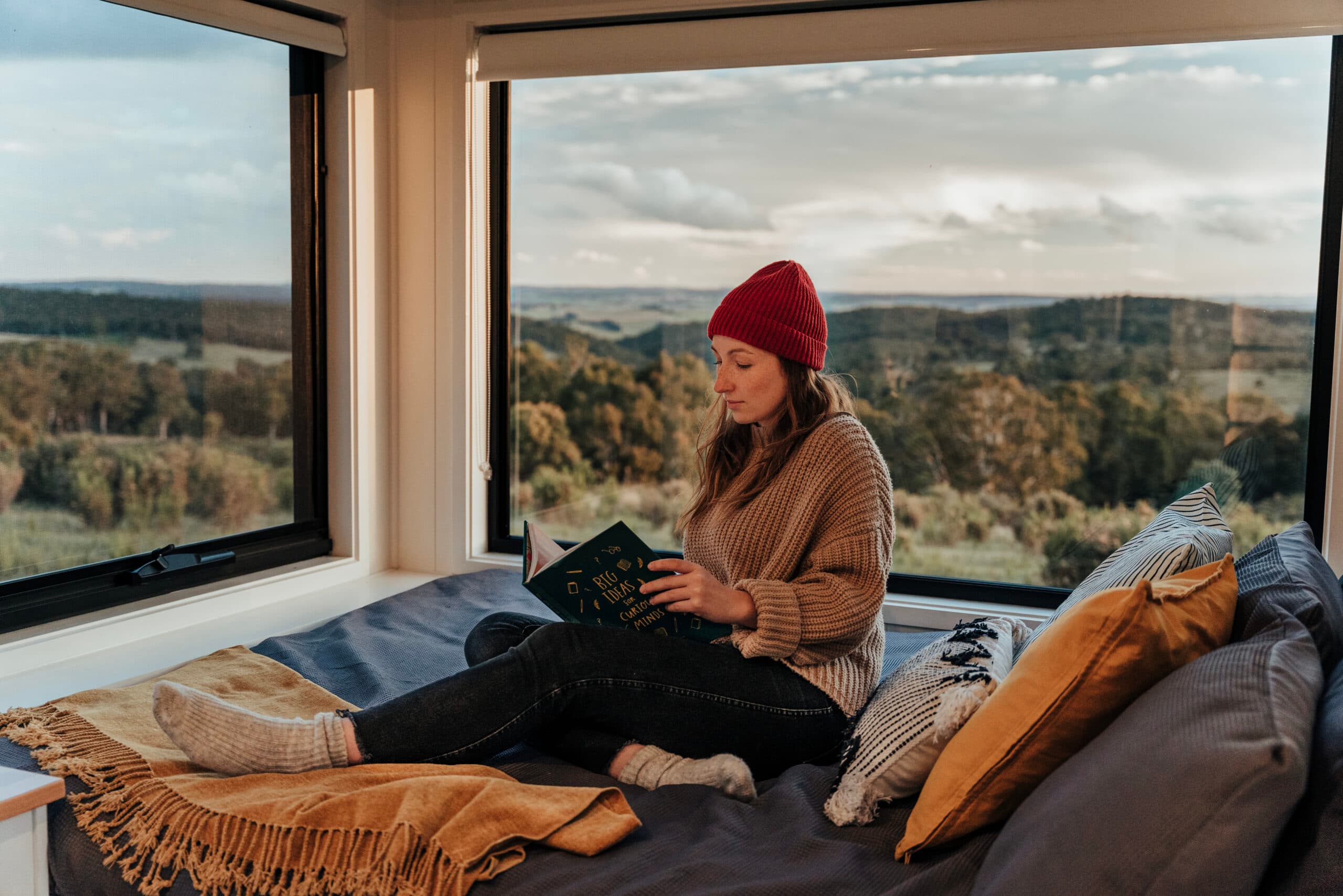 Woman reading in a tiny house with a large window overlooking the Australian bush.