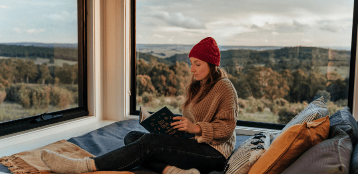 Woman reading in a tiny house with a large window overlooking the Australian bush.